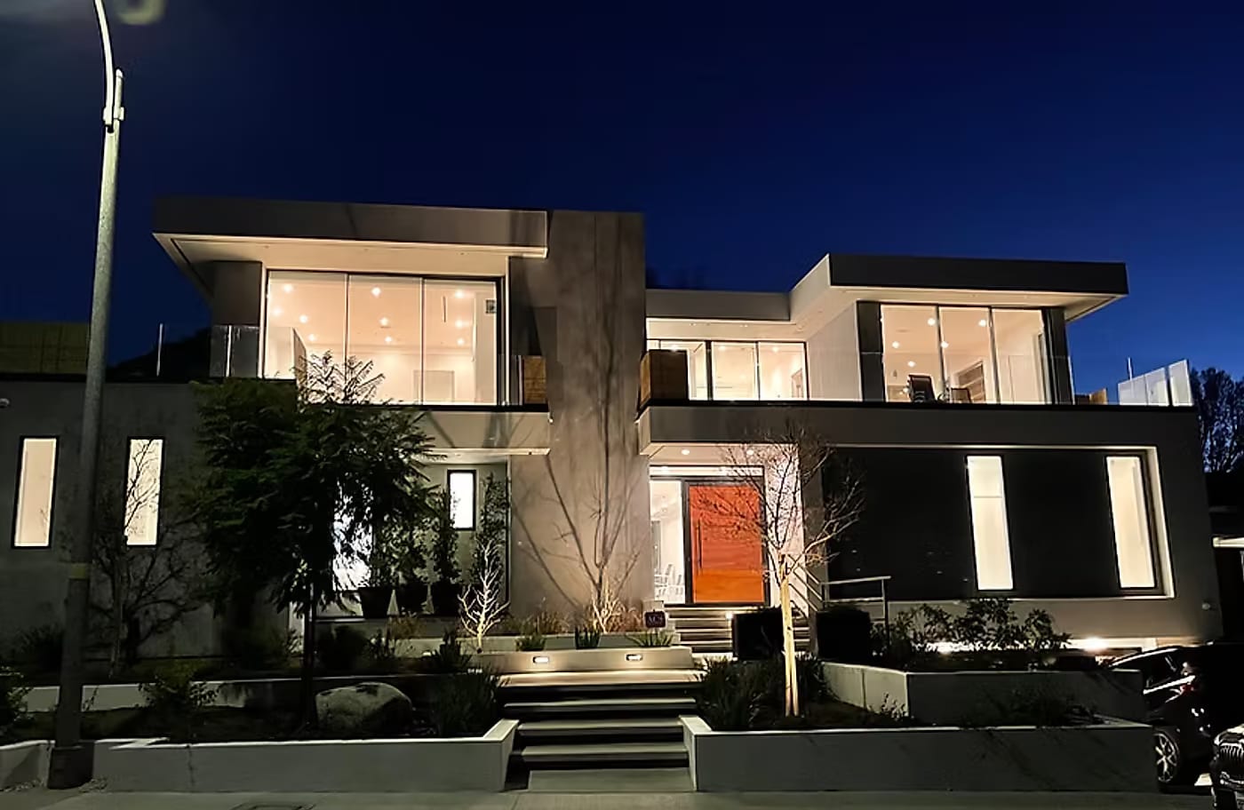The same custom home at dusk with every window lit and a warm timber front door glowing at the top of the entry steps